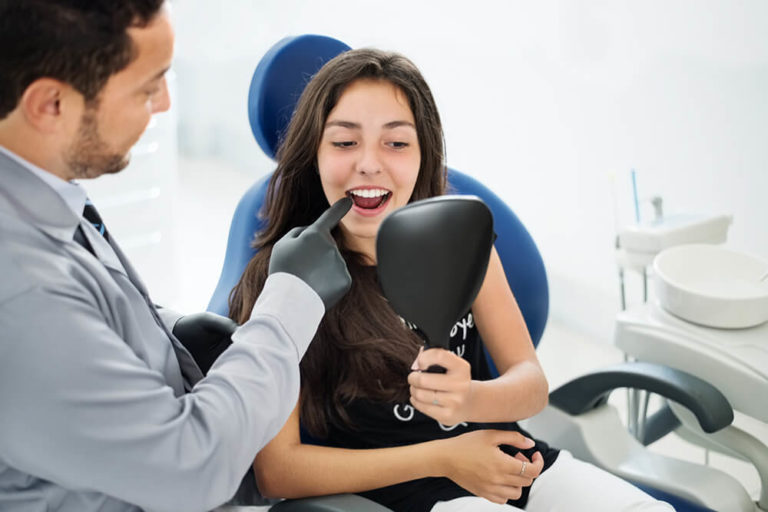 A male dentist points to the teeth of a female dental patient who is seated in a dental chair and holds a mirror to view her teeth