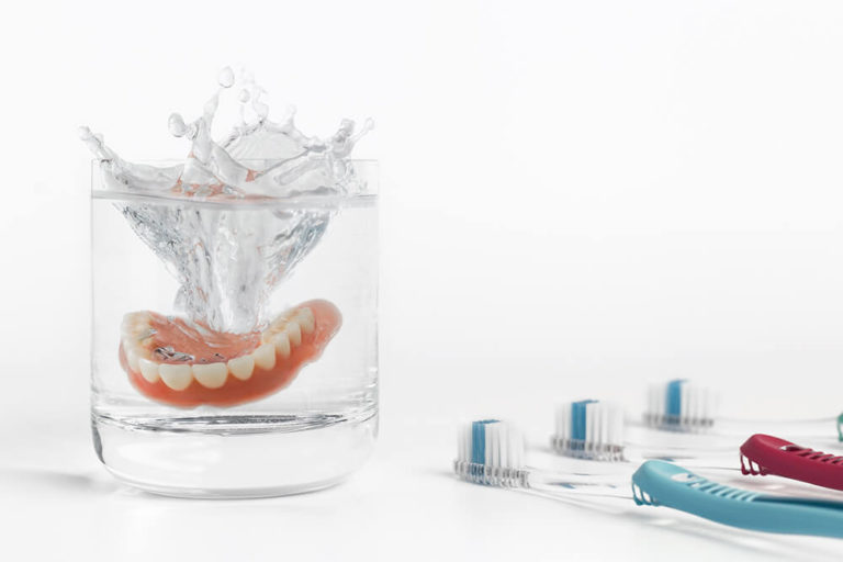 Dentures being dropped into a glass of water with a row of toothbrushes to the right of the glass