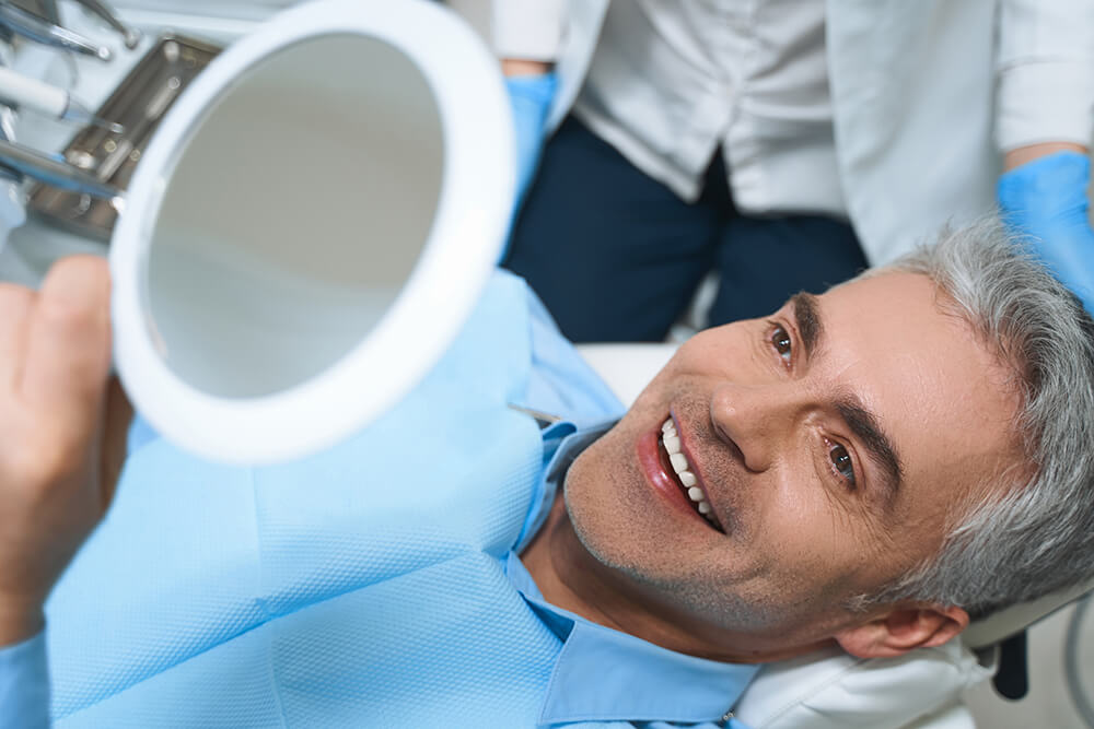 A male dental patient lays back in a dental exam chair and admires his smile in a handheld mirror during a cosmetic dentistry visit